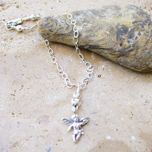 Puede incluir: Un brazalete de tobillo de cadena plateada con un peque&ntilde;o dije de &aacute;ngel plateado. El brazalete est&aacute; sobre una superficie texturizada de color marr&oacute;n claro.
