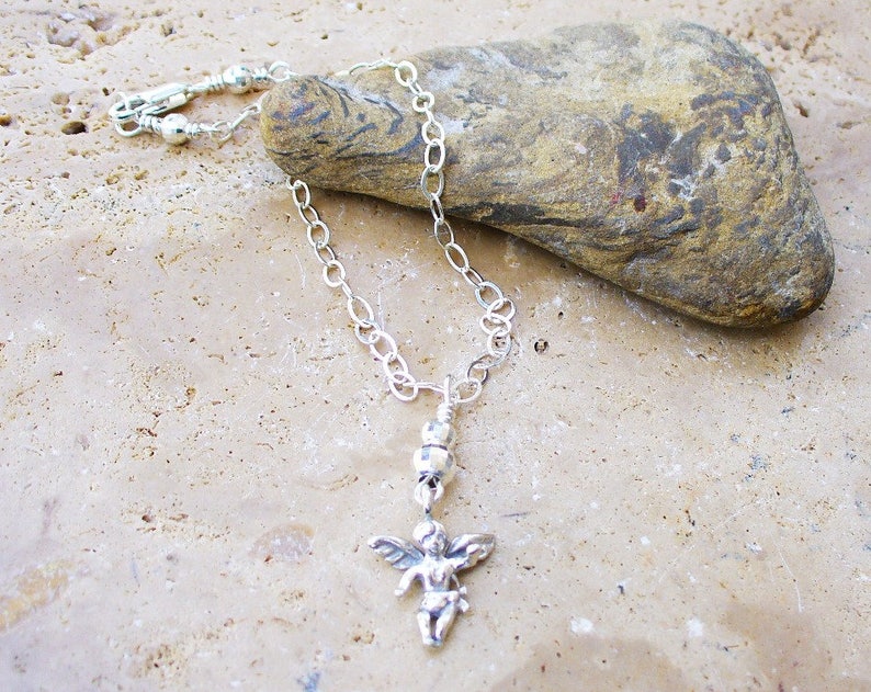 Puede incluir: Un brazalete de tobillo de cadena plateada con un peque&ntilde;o dije de &aacute;ngel plateado. El brazalete est&aacute; sobre una superficie texturizada de color marr&oacute;n claro.