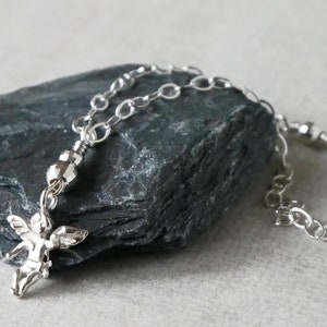 Puede incluir: Un collar de cadena de plata con un peque&ntilde;o dije de &aacute;ngel y una cuenta de plata. El collar est&aacute; sobre una roca gris.