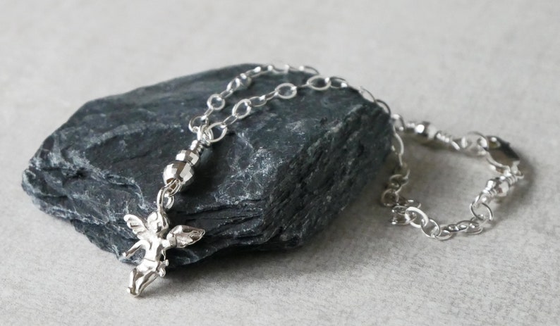 Puede incluir: Un collar de cadena de plata con un peque&ntilde;o dije de &aacute;ngel y una cuenta de plata. El collar est&aacute; sobre una roca gris.