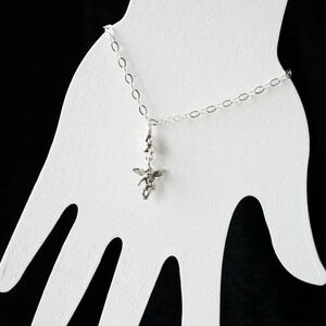 Puede incluir: Un brazalete de cadena de plata con un peque&ntilde;o dije de &aacute;ngel de plata colgando de &eacute;l. El brazalete se muestra en un soporte blanco con forma de mano.