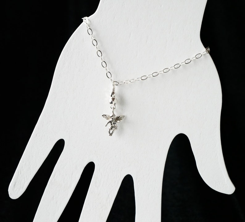 Puede incluir: Un brazalete de cadena de plata con un peque&ntilde;o dije de &aacute;ngel de plata colgando de &eacute;l. El brazalete se muestra en un soporte blanco con forma de mano.