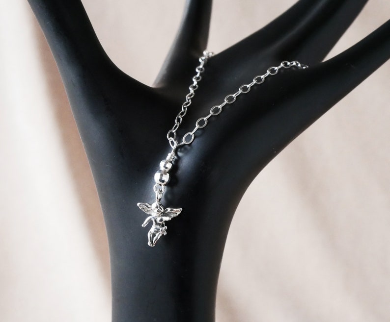 Puede incluir: Un collar de cadena de plata con un peque&ntilde;o dije de &aacute;ngel. La cadena est&aacute; hecha de peque&ntilde;os eslabones redondos.