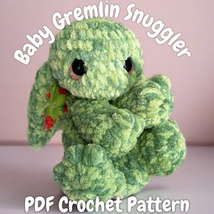 May include: A handmade green and light green crocheted gremlin plush toy with large black eyes and a strawberry-patterned scarf. The text "Baby Gremlin Snuggler" and "PDF Crochet Pattern" are displayed on the image.