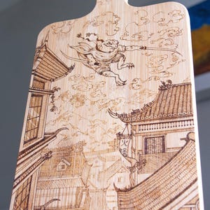 Può includere: Tagliere in legno con manico, con un design pirografato. L'opera raffigura un guerriero rana in un paesaggio urbano in stile giapponese con edifici e nuvole dettagliati. Il tagliere è in legno chiaro.