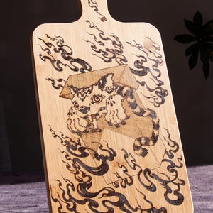 May include: A light-coloured wooden chopping board with a handle, featuring a detailed pyrography design. The design includes a stylized tiger within a geometric shape, surrounded by swirling smoke-like patterns. The board is supported by a black metal stand.