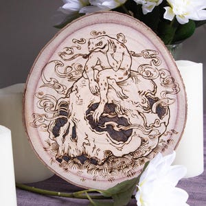 Può includere: Un'opera d'arte rotonda in legno con un design pirografato. L'opera raffigura una creatura simile a una rana seduta su un teschio, circondata da motivi a vortice simili a fumo. Il pezzo è esposto con candele bianche e fiori.