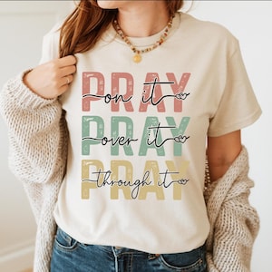 May include: Beige t-shirt featuring the words "PRAY on it, PRAY over it, PRAY through it" in a stacked design. The text is in pink, green, and yellow. The shirt has a crew neck and short sleeves.