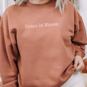 Puede incluir: Una sudadera de color terracota con la frase "Grace in Bloom" en blanco. La sudadera se combina con pantalones blancos. La imagen tiene una estética cálida y otoñal, con calabazas en el fondo.