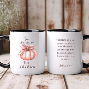 Könnte beinhalten: Zwei weiße Keramikbecher mit schwarzen Griffen und Rändern. Ein Becher zeigt einen Aquarellkürbis mit einer Schleife und dem Text "I am thankful for His Salvation". Der andere Becher hat einen Bibelvers.