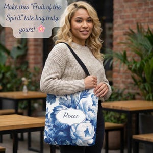 Puede incluir: Un bolso tote blanco con un diseño floral azul y la palabra "Peace" en un marco decorativo. El bolso tiene una correa negra. El bolso lo sostiene una persona.
