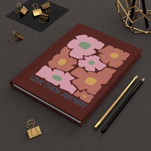 Puede incluir: Un diario marrón con un diseño floral y el texto "LOVE THEM ANYWAY" en la portada.