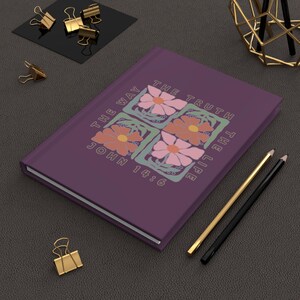 Puede incluir: Un diario morado con el texto "THE WAY THE TRUTH THE LIFE JOHN 14:6" y diseños florales. También se ven lápices dorados y negros y clips dorados.