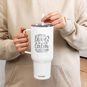 Puede incluir: Vaso blanco con asa y tapa, con el texto "With His love, He will calm all your fears" y el versículo bíblico Sofonías 3:17. Incluye una pajita blanca. La marca "koozie" está impresa en la parte inferior.