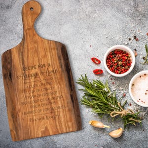 Puede incluir: Una tabla de cortar de madera con una receta para un hogar feliz grabada en ella. La receta incluye ingredientes como el amor, la lealtad, la paciencia, la risa, la esperanza y el perdón. La tabla está rodeada de ramitas de romero, dientes de ajo y granos de pimienta roja.