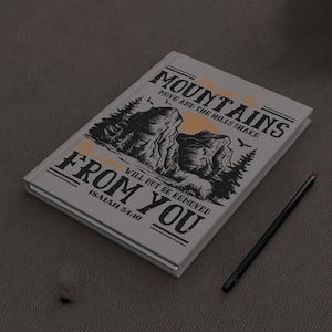 Puede incluir: Un cuaderno gris con una ilustración en blanco y negro de montañas y árboles. El texto "Las montañas no serán quitadas de ti" está impreso en la portada, junto con la referencia bíblica "Isaías 54:10".