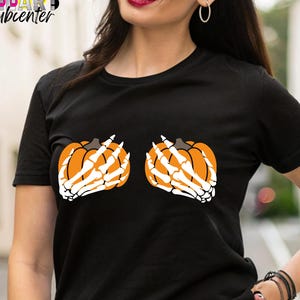 May include: Black t-shirt featuring a Halloween-themed design. The shirt displays two orange pumpkins with white skeleton hands. The design is centered on the chest. The shirt has short sleeves and a crew neck.