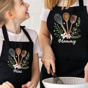 May include: Black aprons with a floral design and the words "Mommy" and "Mini" printed in white. The aprons feature illustrations of a whisk, spoon, and spatula. The aprons are worn by a woman and a child.