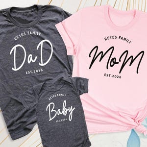 May include: Three matching family shirts and a onesie. The dark grey t-shirt says "Dad" in white script, the pink t-shirt says "Mom" in black script, and the dark grey onesie says "Baby" in white script. All items have "Reyes Family Est. 2026" printed above or below the main text.