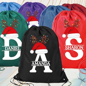 May include: A collection of drawstring bags in various colors, including green, black, red, and blue. Each bag features a festive design with a Santa hat, reindeer antlers, and a personalized name in white. The bags are ideal for Christmas gifts.