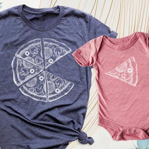 May include: A navy blue t-shirt and a dusty rose onesie. Both garments feature a white pizza slice graphic. The t-shirt has a large pizza slice design, while the onesie has a single slice. The pizza design includes toppings.
