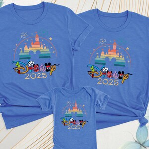 May include: Blue t-shirts and a onesie featuring a colorful castle design with the text "Disney 2025". The design includes Mickey and Minnie Mouse characters. The shirts are short-sleeved.