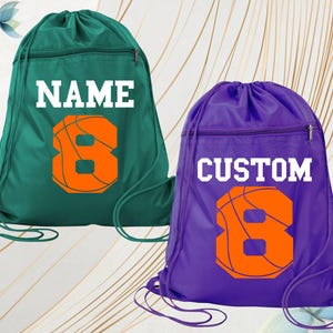 May include: Two drawstring sports bags, one teal and one purple. Each bag features a basketball graphic with the number 8 in orange. The teal bag says "NAME" in white, and the purple bag says "CUSTOM" in white.