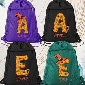 May include: Four drawstring backpacks in purple, black, and teal. Each bag features a large letter with a Halloween theme, including a witch hat and spiderwebs. Names are printed below the letters.