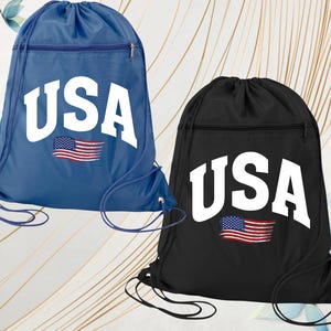 May include: Two drawstring bags, one blue and one black, each featuring the word "USA" in bold white letters. Below the text is a graphic of the American flag. Both bags have a zippered pocket and drawstring closure.