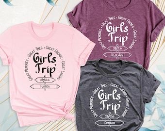 May include: Three t-shirts in light pink, dark purple, and charcoal gray. Each features a circular graphic with the text Girls Trip, Great Times, Great Friends, Great Laughs, Great Memories, 2026, and a location: Florida, Key West, or Branson.