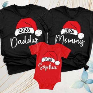 May include: Two black short-sleeve t-shirts and one red short-sleeve baby bodysuit. Each garment features a red Santa hat graphic with the year 2026 printed on the white trim. The shirts say Daddy and Mommy, while the bodysuit says Sophia.