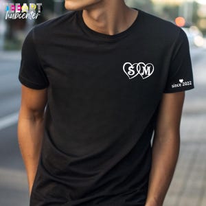May include: Black t-shirt with white heart-shaped graphic featuring the letters "S" and "M". The right sleeve has the text "since 2022". The shirt is a classic crew neck style.