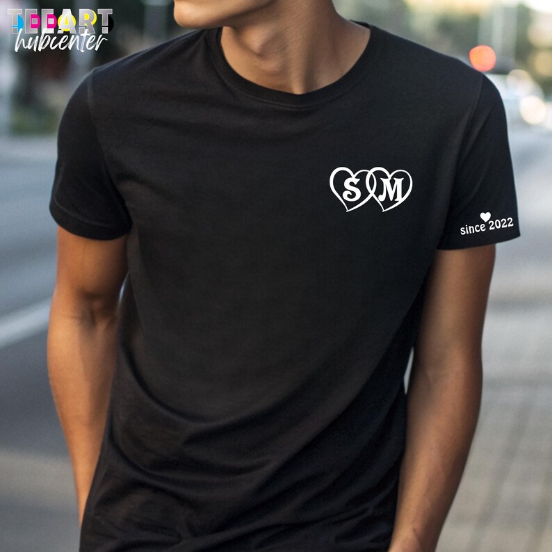 May include: Black t-shirt with white heart-shaped graphic featuring the letters "S" and "M". The right sleeve has the text "since 2022". The shirt is a classic crew neck style.