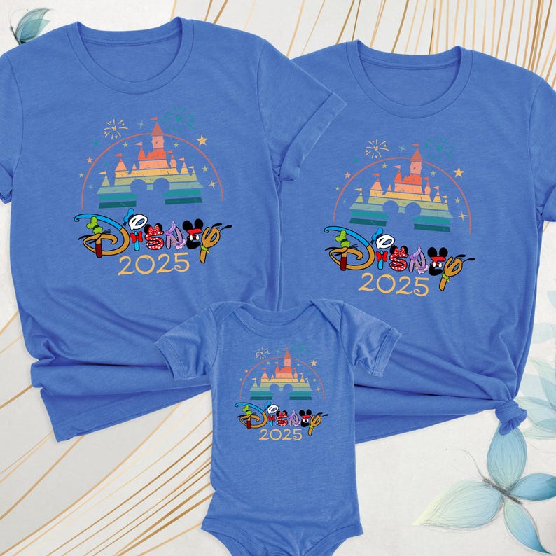 May include: Blue t-shirts and a onesie featuring a colorful castle design with the text "Disney 2025". The design includes Mickey and Minnie Mouse characters. The shirts are short-sleeved.