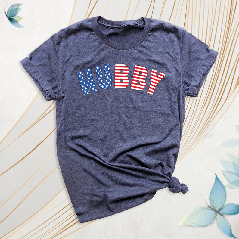 Puede incluir: Una camiseta azul marino con la palabra "HUBBY" en rojo y azul, con letras que recuerdan la bandera estadounidense. La camiseta est&aacute; anudada en la parte inferior y tiene mangas cortas.