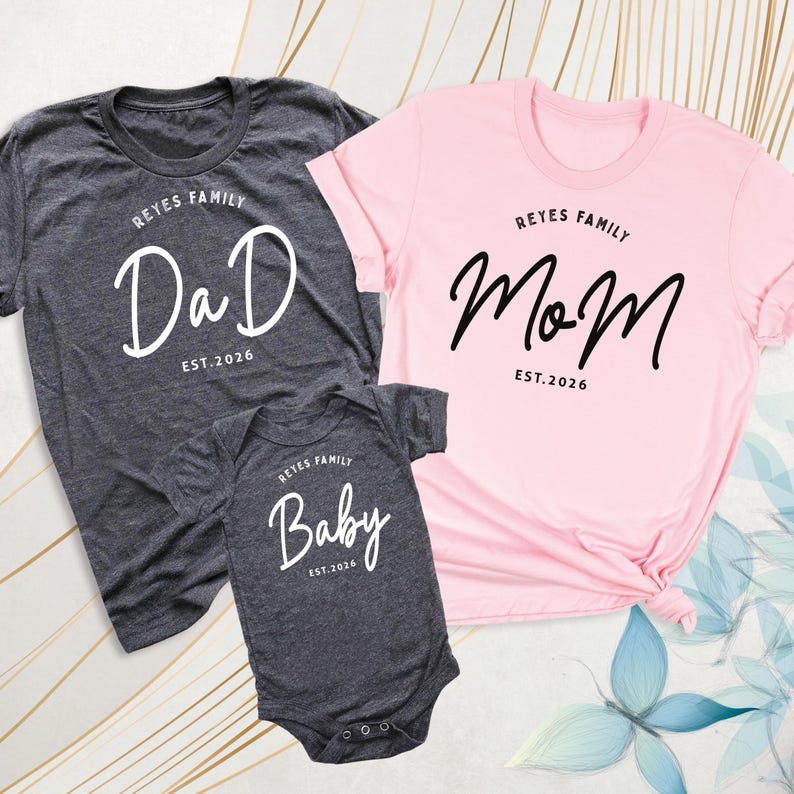 May include: Three matching family shirts and a onesie. The dark grey t-shirt says "Dad" in white script, the pink t-shirt says "Mom" in black script, and the dark grey onesie says "Baby" in white script. All items have "Reyes Family Est. 2026" printed above or below the main text.