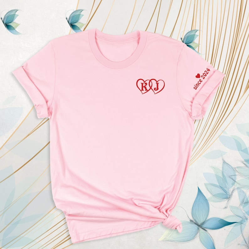 May include: A light pink t-shirt with rolled-up sleeves. The shirt features two red hearts with the letters "R" and "J" inside. The text "since 2024" is printed on the sleeve.