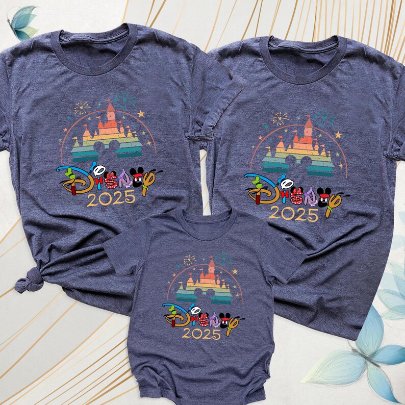 May include: Three blue t-shirts and a onesie with a colorful graphic. The graphic includes a castle, fireworks, and the text "Disney 2025" in a stylized font. The shirts are short-sleeved.