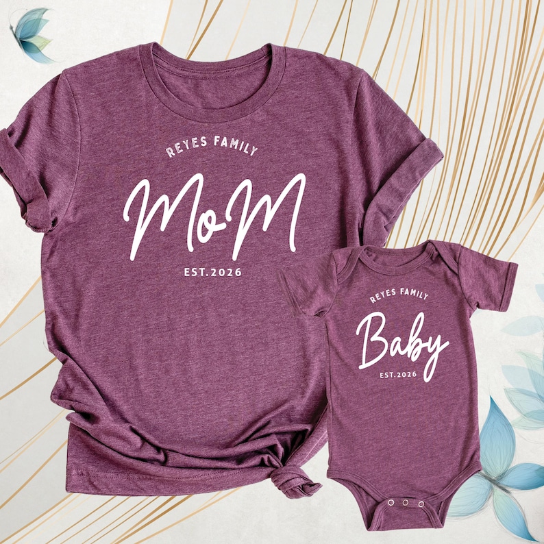 May include: Burgundy t-shirt and baby bodysuit set. The adult shirt features the word "Mom" with "Reyes Family" above and "Est. 2026" below. The baby bodysuit says "Baby" with "Reyes Family" above and "Est. 2026" below.