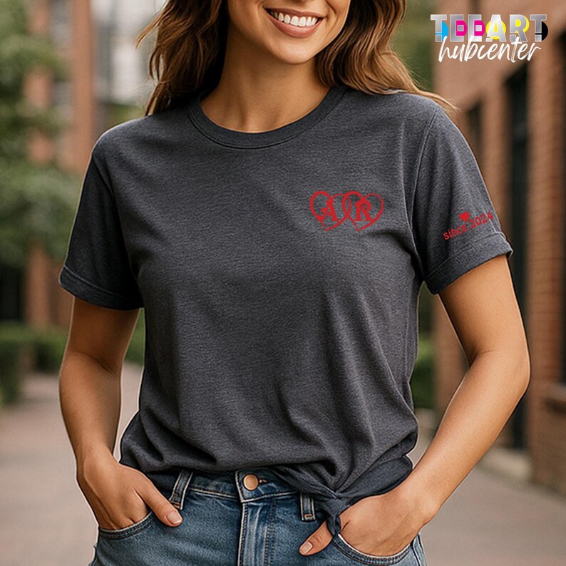 May include: Dark gray t-shirt with a red logo on the chest and the text "Since 2024" on the sleeve. The shirt has a crew neck and short sleeves. The model is wearing blue jeans.