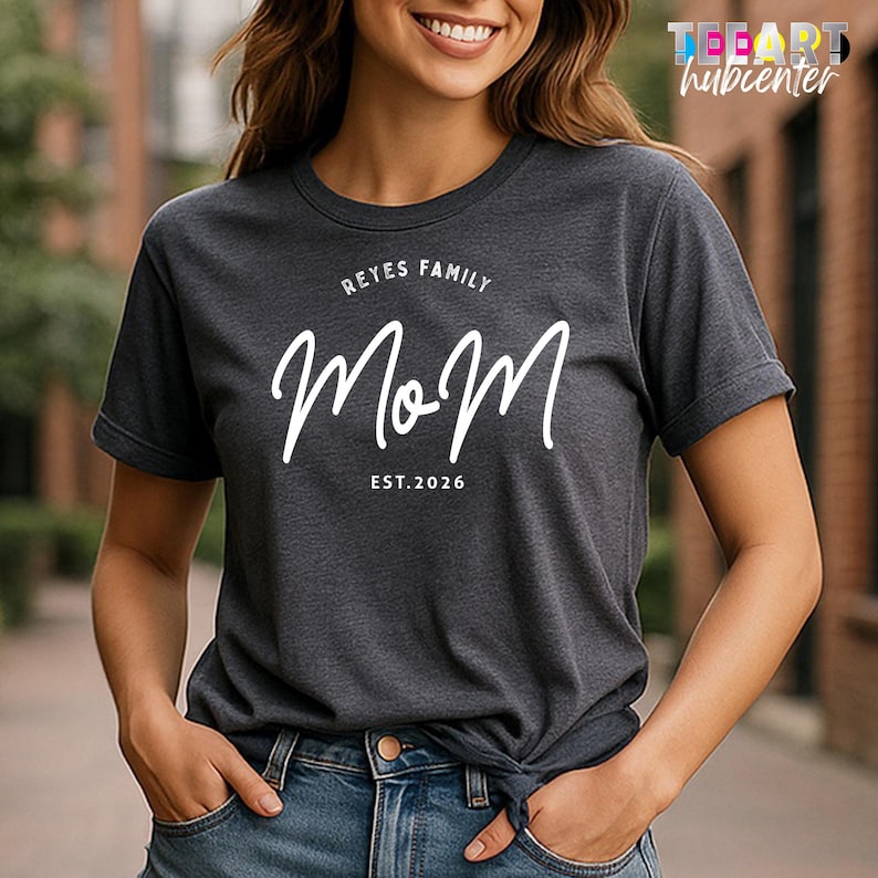 May include: Dark grey t-shirt featuring the text "Reyes Family Mom EST. 2026" in white script. The t-shirt has a crew neck and short sleeves. The fabric appears soft.