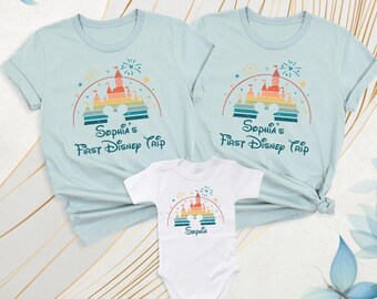 May include: Two light blue short-sleeve t-shirts and one white short-sleeve baby bodysuit. Each garment features a colorful graphic of a castle, a rainbow, fireworks, and the text "Sophia's First Disney Trip" or "Sophia".