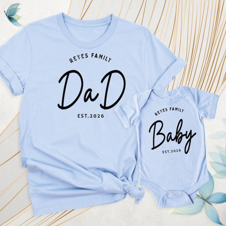 May include: Pale blue matching shirts. One is a t-shirt with the text "REYES FAMILY DaD EST.2026" in black script. The other is a baby onesie with the text "REYES FAMILY Baby EST.2026" in black script.