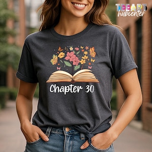 May include: Dark gray t-shirt featuring a graphic of an open book with flowers and butterflies. The text "Chapter 30" is printed below the book. The shirt is a crew neck style.