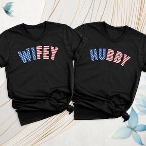 Puede incluir: Dos camisetas negras con las palabras "WIFEY" y "HUBBY" impresas. El texto est&aacute; en rayas rojas y blancas con un patr&oacute;n de estrellas azules. Las camisetas son probablemente para parejas.