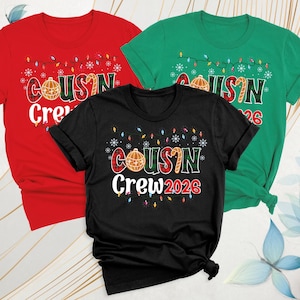 May include: Three short-sleeve t-shirts in red, green, and black. Each shirt features a graphic with the text "COUSIN Crew 2026" in a plaid font, decorated with a Christmas ornament, a candy cane, string lights, and snowflakes.