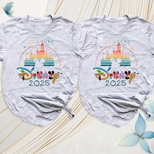 May include: Two light gray t-shirts with a colorful graphic featuring a castle, fireworks, and the word "Disney" in a stylized font. The year "2025" is printed below the logo. The shirts are folded at the bottom.