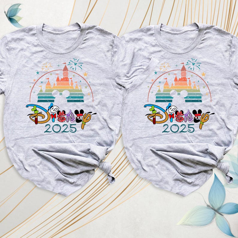 May include: Two light gray t-shirts with a colorful graphic featuring a castle, fireworks, and the word "Disney" in a stylized font. The year "2025" is printed below the logo. The shirts are folded at the bottom.