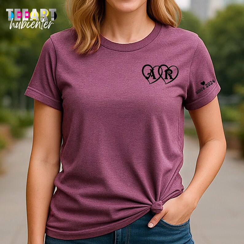 May include: A maroon t-shirt with a round neck and short sleeves. The shirt features a design of two intertwined hearts with the letters "A" and "R" inside. The text "since 2024" is printed on the sleeve.