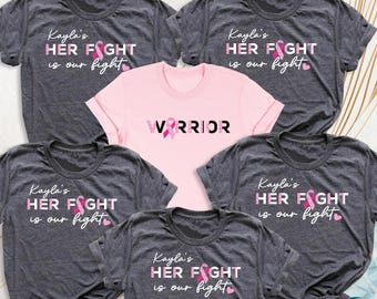 May include: A collection of gray and pink t-shirts. The gray shirts display the text "Kayla's HER FIGHT is our fight" with a pink ribbon graphic. A pink shirt in the center reads "WARRIOR" with a pink ribbon. The shirts are short-sleeved.